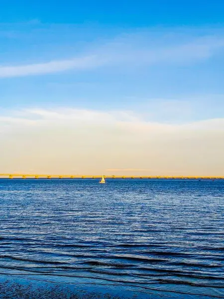 Denizde küçük bir yelkenli. Arka planda deniz suyunu aydınlatan bir köprü var.