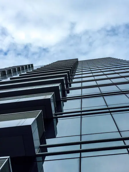 Parçalı bulutlu mavi gökyüzüne karşı yansıtıcı cam paneller ile çağdaş bir gökdelenin üst görünümü, mimari tasarım ve kentsel gelişim sergiliyor.