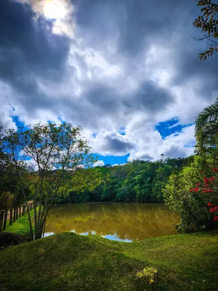 Dramatik, bulutlu bir gökyüzünün altında sakin bir göl manzarası, kıyı boyunca canlı yeşil yapraklar ve kısmen görülebilir bir güneş bulutların arasından gözetliyor..