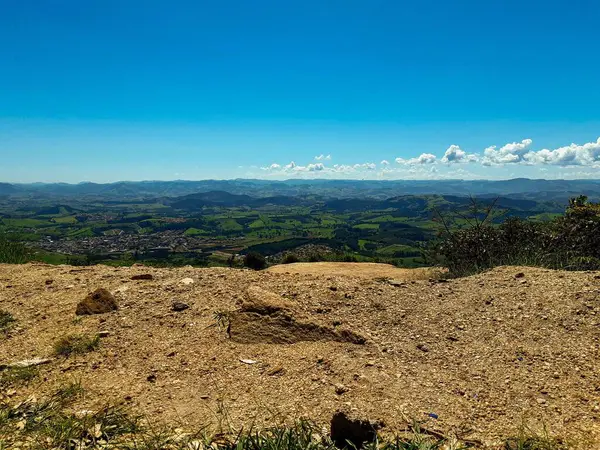 Ön planda kurak kayalık arazi ile geniş doğal manzara, berrak mavi bir gökyüzü altında yemyeşil tepelere dönüşüyor tüylü beyaz bulutlar, huzuru çağrıştırıyor.