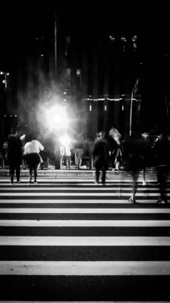 Parlak ışıklı yaya geçidini geçen insanların büyük bir şehirde canlı bir gece görüntüsü. Hareketli figürler, şehir yaşamının enerjisini yakalayan bir aciliyet ve dinamizm duygusu yaratıyor..