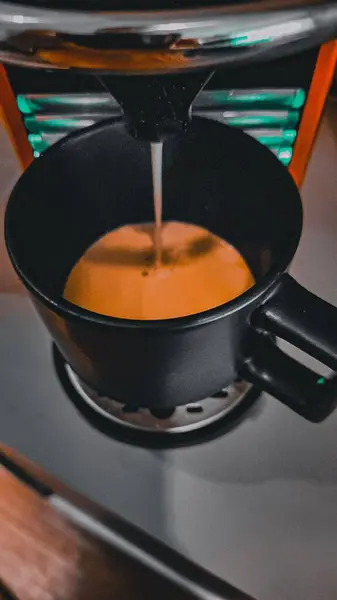 Görünür buharı ve rahat bir hissi olan bir makineden taze demlenen siyah bir fincan kahve. Detaylar kahvenin zengin rengini ve arka plandaki modern makineyi vurguluyor..