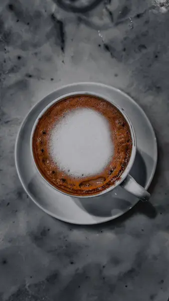 Beyaz bir fincan kahve mermer bir yüzeye oturur. Kahvenin tepesinde bol kremalı köpük tabakası var. Yumuşak, kadife gibi bir dokusu var..