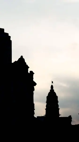 Şafak vakti yumuşak bir gökyüzüne karşı tarihi binaların dramatik siluetleri. Mimari biçimler, barış ve kentsel nostalji hissi uyandıran parlak arka plana karşı çıkıyor. Süslü detaylar yükselen güneş tarafından geliştirilir..