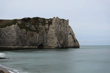 Etretat ikonik kayalıkları gri bulutların altında hareketsiz ve görkemli duruyor, sakinlik hissi uyandırıyor..