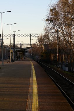 Bir demiryolu platformunun canlı bir görüntüsü, çerçevenin kenarında uzanan parlak sarı bir güvenlik çizgisi, hattın çevreye karşı kontrastını vurguluyor. Uzakta, demiryolu kızılötesi eşliğinde beton bir yolcu sığınağı görülebilir.