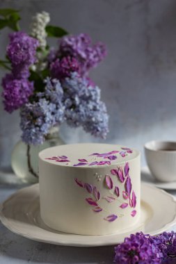 Elegant cake with floral buttercream decorations and lilac bouquet in soft natural light, perfect for celebrations and artistic photography concepts clipart