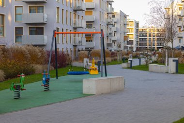 Güneşli bir akşamda, apartman binaları ve manzaralı yeşilliklerle çevrili çocuk parkı olan modern bir konut avlusu. Kentsel yaşam, aile yaşam tarzı ve açık hava eğlence alanları