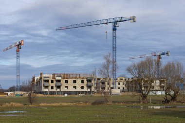 Beton duvarları, iskelesi ve bulutlu gökyüzüne karşı yüksek vinçleri olan modern bir yerleşim yerinin inşaatı devam ediyor.