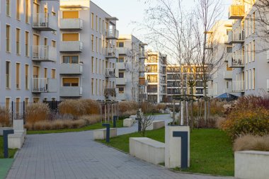 Yeşil avlu, asfaltlı yollar, yeşillik ve kentsel bir ortamda hafif güneş ışığıyla aydınlatılan modern konut kompleksi.