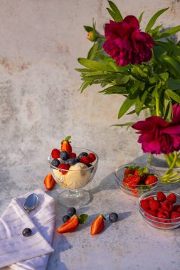 Zarif bir yaz tatlısı vanilyalı dondurma ve taze böğürtlenler, bir cam kasede servis edilmiş arka planda kırmızı şakayık buketinin yanında. Yaz ziyafetleri ve çiçek dekoru