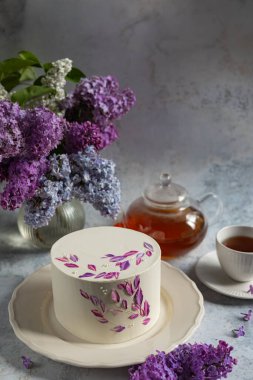 Seramik bir tabakta çiçekli mor süslemeli zarif beyaz pasta çiçek açan leylaklar, bir çaydanlık ve bir fincan çayla çevrili.
