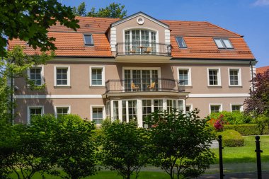 Zarif lüks villa bej cepheli, kırmızı fayanslı çatı ve yemyeşil bahçe ve ağaçlarla çevrili balkonlar. Modern mimari, lüks yaşam ve huzurlu konut