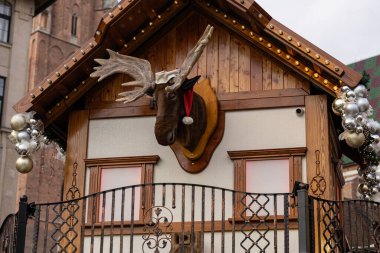 Festive wooden chalet at the Wroclaw Christmas Market featuring a decorative moose head in a Santa hat, adorned with sparkling silver ornaments, fairy lights, and intricate wrought iron railing clipart