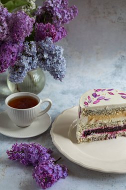 Beyaz bir tabakta kremalı ve böğürtlenli haşhaş tohumu keki, yanında bir fincan çay ve pastel arka planda bir buket taze leylak çiçeği.