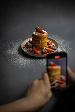 Person photographing a plate of fluffy Japanese pancakes with fresh berries, powdered sugar, and syrup using a smartphone. Food photography, culinary presentation, and social media content creation clipart