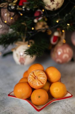 Ön planda soyulmuş bir meyve bulunan taze mandalinalar güzel dekore edilmiş bir Noel ağacının yanına yerleştirilmiş. Bayram neşesi, mevsimlik meyve ve bayram gelenekleri