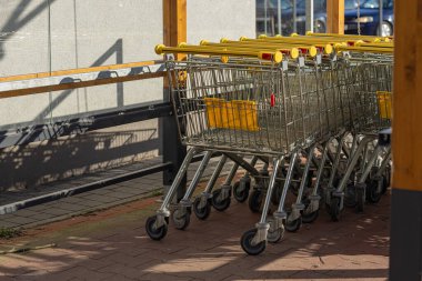 Sarı saplı metal alışveriş arabaları süpermarketin yakınındaki bir araba deposunda güneş ışığıyla aydınlatılıyor. Perakende kolaylığı, organizasyon ve alışveriş merkezleri