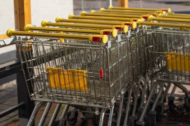 Sarı saplı metal alışveriş arabaları güneşli bir günde koruyucu bir gölgeliğin altında süpermarketin önünde düzgünce dizilmiş. Perakende satış, market alışverişi, kolaylık ve modern tüketici alışkanlıkları