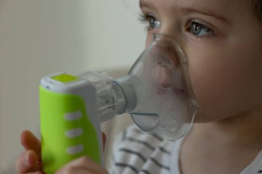 Evde solunum tedavisi için yeşil saplı nebulizör kullanan parlak gözlü bir çocuğun yakın çekimi. Pediatrik astım, solunum terapisi ve solunum desteği için tıbbi ekipman.