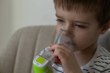 Evdeki koltukta otururken nebulizör spreyi kullanan kısa kahverengi saçlı küçük bir çocuk. Solunum terapisi, astım tedavisi, sağlık hizmetleri ve solunum desteği için pediatrik tıbbi ekipmanlar