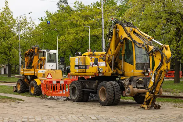 Wroclaw, Poland- 24 Nisan 2025: Güvenlik bariyerleri ve ağaçları olan bir caddenin bakım çalışmaları sırasında tramvay hattının yakınına park edilmiş iki sarı inşaat kazıcısı. Kentsel altyapı ve onarım