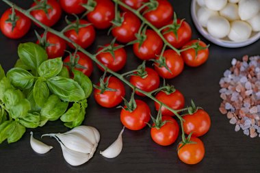 Fesleğen yapraklı taze kiraz domatesleri, sarımsak karanfilleri, Himalaya pembe tuzu ve siyah ahşap masada mozzarella topları. Ev yapımı salata hazırlamak için doğal malzemeler.