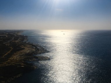 Sakin mavi sulara bakan kayalık bir sahil yolundan, parlak güneş ışığı engin denizde parıldıyor ufka yakın, bulutsuz bir gökyüzü ve kıyı çalılarının altında sürüklenen küçük bir tekne gibi..