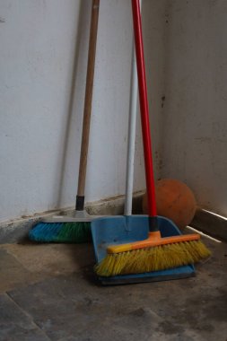 Kırmızı saplı süpürge ile mavi bir faraş üzerinde dinlenen sarı tüylü süpürge de dahil olmak üzere temizlik araçlarının yer aldığı bir köy köşesi, yeşil tüylü başka bir süpürgeyle birlikte. Arka planda sönük turuncu bir basketbol topu yıpranmış, desenli bir w 'ye yaslanıyor.