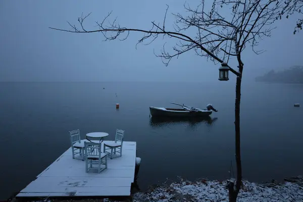 Sakin bir kış sahnesi, sakin bir göle bakan, sandalyeli ve masalı karla kaplı bir rıhtım. Yakınlarda tek bir tekne yüzüyor ve içinde fener olan çıplak bir ağaç, sakin atmosfere katkı sağlıyor. Sessiz tonlar ve sisli arka plan huzurlu bir çağrışım yapıyor, 
