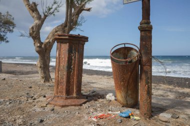Paslı bir çöp tenekesi ve bir kolon birbirine bağlanmış bir iple kumlu ve kayalık bir sahilde çıplak bir ağaç gövdesinin yanında duruyordu. Çöp, çöp kutusunun etrafına saçılmış. Arka planda, okyanus dalgaları kıyıya doğru yuvarlanırken ufukta...