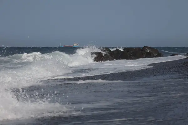 Görüntü dalgaların kıyı şeridindeki sivri siyah kayalara çarptığını ve ufukta bir kargo gemisinin göründüğünü gösteriyor. Dalgalar kayalara çarptıkça su sıçratır ve köpürür.