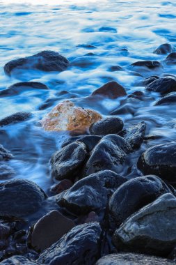 Bu yakın plan, bir kıyı şeridi veya nehir kıyısı boyunca kısmen akan suya batmış koyu renkli kayaları tasvir eder. Uzun pozlama tekniği, su üzerinde ipeksi bir etki yaratır. Kayaların keskin detaylarıyla çelişir..