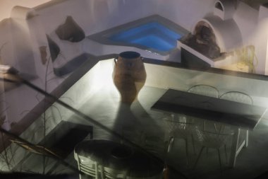 Büyük bir seramik vazo, bir yemek masası ve küçük ışıklı bir havuzu olan bir açık hava avlusu içeren gerçeküstü, katmanlı bir gece sahnesi. Hareket bulanıklığı ve yansımalar soyut ve rüya gibi bir etki yaratarak Akdeniz arşivinin elementlerini harmanlıyor