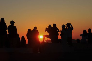 Siluetli figürler gün batımını izlemek için bir çıkıntı boyunca toplanırlar. Şekilleri alacakaranlığın sıcacık ışığıyla harmanlanır. Sıradan pozlar, şapkalar ve rahat duruşlar akşam gökyüzünün güzelliği için ortak bir takdiri yansıtır.