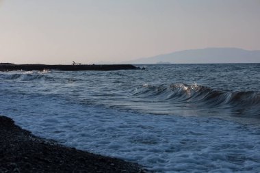 Issız bir iskelede, çakıl taşlarıyla kaplı bir kıyıda dalgalar yuvarlanan tek bir sandalye.