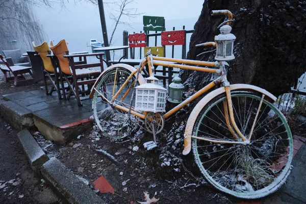Karla kaplı, fenerli sarı bir bisiklet, sakin bir kış sabahı boyunca renkli sandalyelerin yanında ve göl kenarında bir verandada dinlenir..