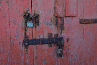 Paslı bir zincir ve eski kırmızı ahşap kapıyı koruyan soluk mavi bir asma kilit. Soyulmuş boya ve aşınmış metal desenli, eski ve terk edilmiş bir görünüş yaratır..