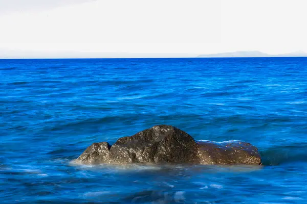 Derin mavi okyanusun pürüzsüz, akışkan hareketinden koyu, dokulu bir kaya çıkar. Etrafında hafif dalgalar dalgalanır, sükunet ve hareket hissi yaratır.