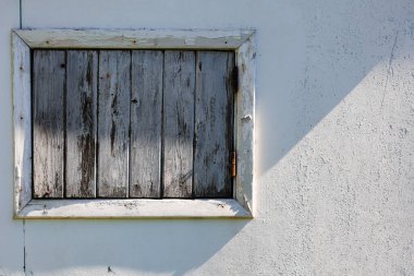 Beyaz desenli bir duvarda boyası soyulan eski ahşap bir pencere perdesi güneş ışığında güçlü bir çapraz gölge oluşturur.
