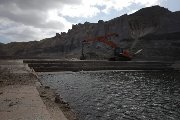 Turuncu kazıcı durgun bir su kütlesinin yakınındaki engebeli uçurumların ortasında duruyor..