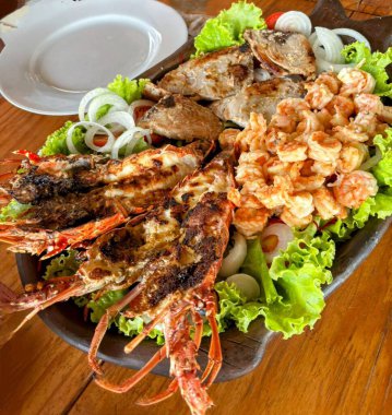 Lezzetli, ızgara ıstakoz, karides ve balıklı ızgara deniz ürünleri tabağı taze marul yatağında servis edilir ve dilimlenmiş soğan ve domatesle süslenir..