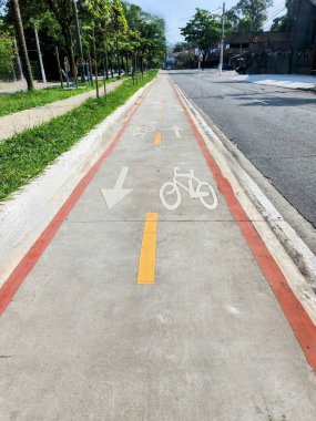Çimenli kaldırım ve bulvarın kenarlarında seyrek ağaçlar ve binalar olan cadde arasına yerleştirilmiş, net yönlü ok işaretleri ve bisiklet sembolleriyle sakin, boş bir şehir bisikleti yolu..