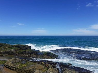Güneşli bir günde açık mavi bir gökyüzü ve geniş bir okyanusu tasvir eden, kayalara çarpan kayalık bir kıyı şeridinin panoramik görüntüsü..