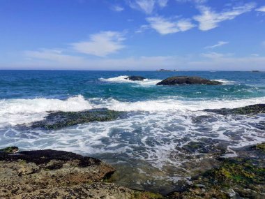 Güneşli mavi bir gökyüzünün altında yumuşak dalgaların dalgalandığı kayalık bir kıyı şeridinin panoramik görüntüsü. Görüntü, kıyı manzarasının dingin güzelliğini ve doğal sükunetini yansıtıyor..