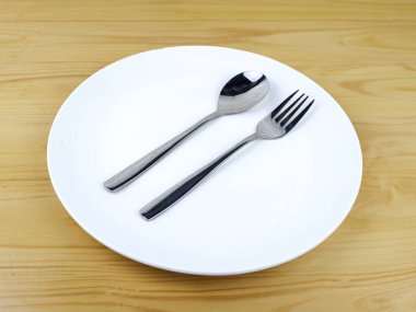 Tahta masadaki boş beyaz tabakta bir kaşık ve bir çatal.