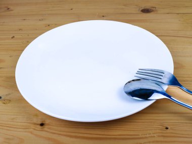 Tahta masadaki boş beyaz tabakta bir kaşık ve bir çatal.