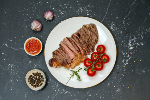 A perfectly grilled ribeye steak, sliced to reveal a juicy medium-rare center, served on a white plate. Accompanied by fresh cherry tomatoes, garlic, peppercorns, and a side of rich red sauce. The dish is presented on a dark marble surface.