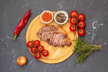 Altın hamurlu pirzola, yuvarlak bir ahşap kesme tahtasının üzerindedir. Taze vişneli domatesler, kırmızı biber, soğan, kekik, kaba tuz, karışık karabiber ve baharatlı kırmızı sos ile çevrili. Koyu mermer bir arka planda gurme yemeği