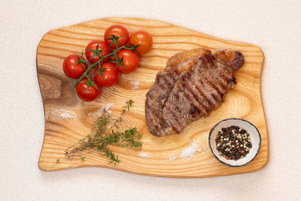 A grilled ribeye steak with a golden-brown crust rests on a rustic wooden cutting board. Accompanied by cherry tomatoes, coarse salt, a mix of black and white peppercorns, and fragrant thyme sprigs. A gourmet meal presented in a minimalist style